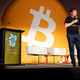 Alex Mashinsky stands on a stage with a microphone in his hand wearing a shirt reading "Celsius." Behind him are several empty chairs and a giant Bitcoin symbol.